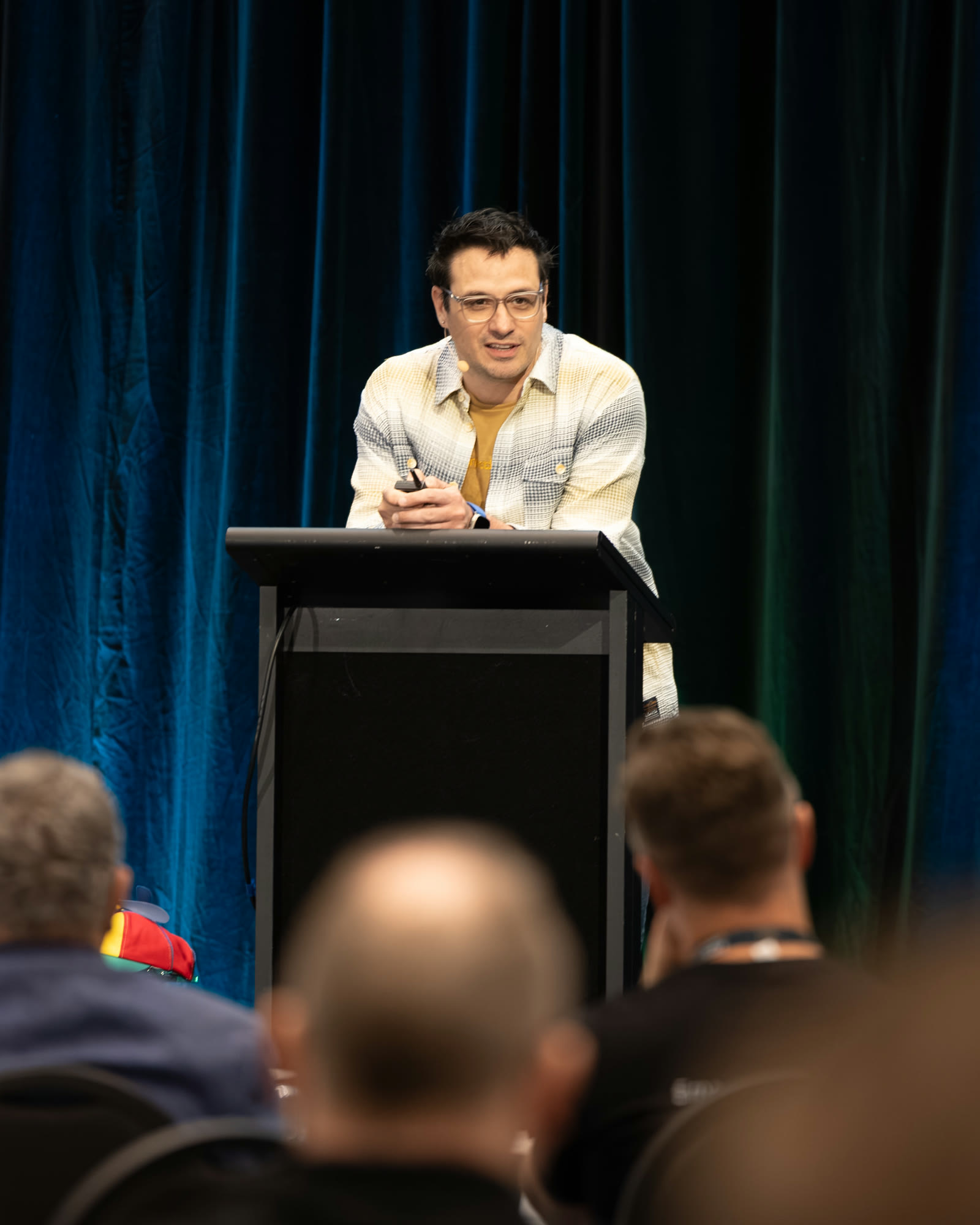 Diego Nievas speaking from a lectern at a conference, leaning forward mid-sentence, blurred audience heads in the foreground.