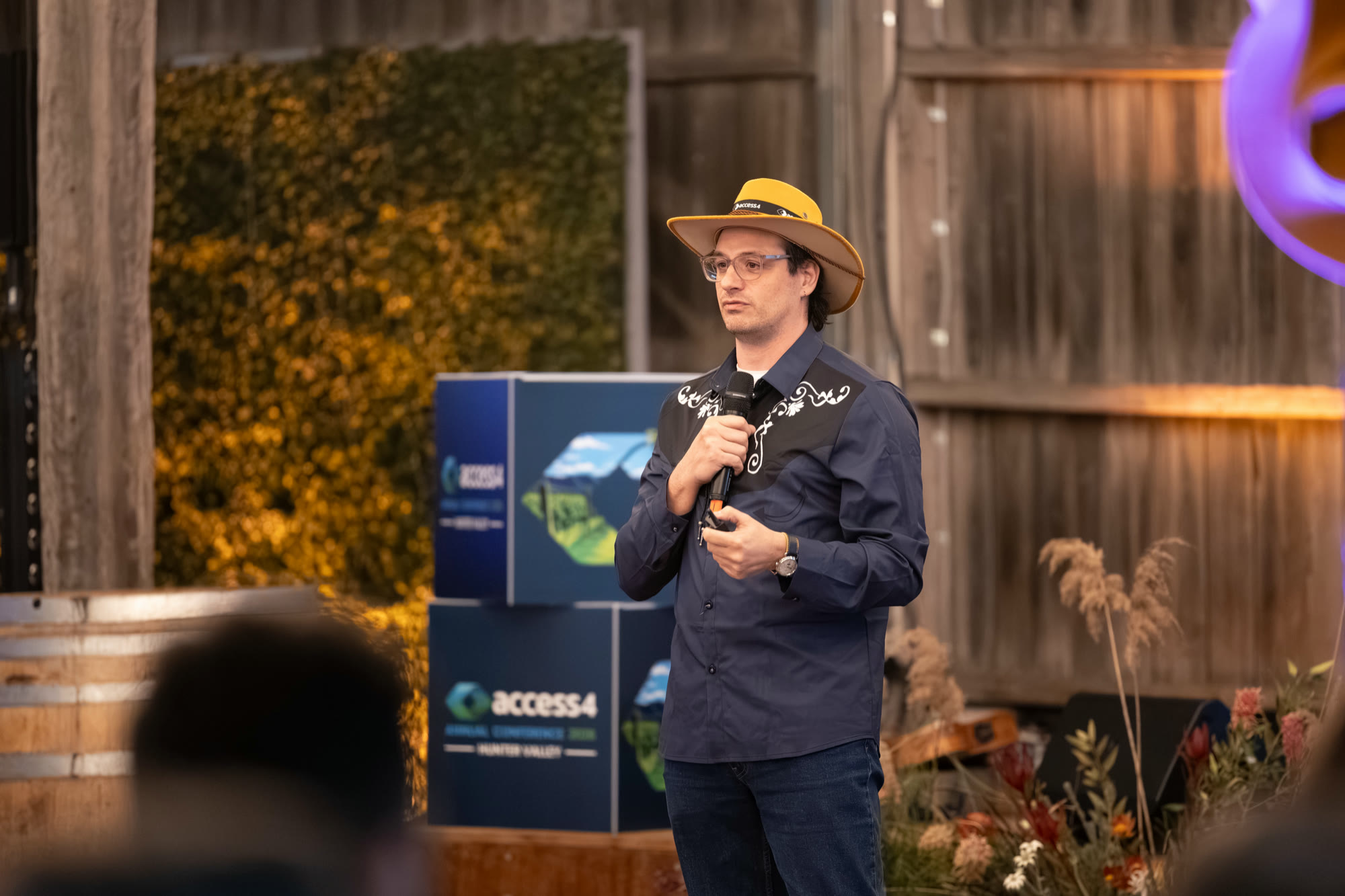 Diego Nievas at the gala in a dark shirt and wide-brimmed hat, holding a microphone in front of a wall of yellow flowers.