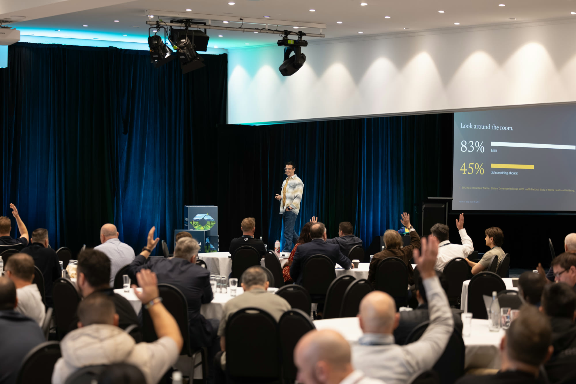 The conference room mid-keynote with hands raised across the audience, the slide behind showing two figures — 83% and 45% — as horizontal bars.